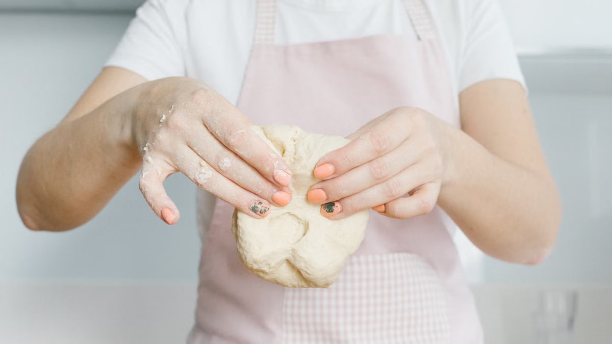 A woman wearing a pink apron kneads dough in a bright, modern kitchen.